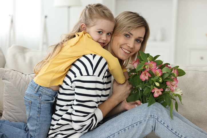 Little daughter congratulating her mom with Mother`s Day at home. Woman holding bouquet of alstroemeria flowers