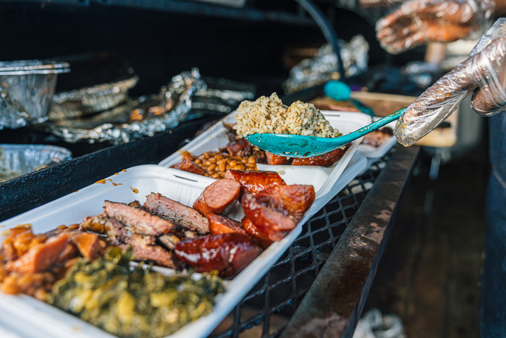 Catering Chef adds a large generous scoop of Dirty Rice to a Compostabale Takeout Container full of Smoked BBQ Beans, Sausage, Chicken Thighs & Drumsticks at a Food Truck or Traveling Smoker Catering Company