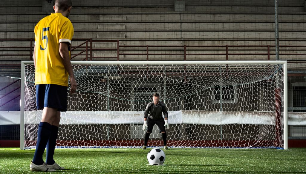 Two soccer players on a field, one in a yellow jersey standing in front of a goal net.