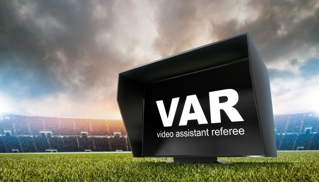 A large black video assistant referee (VAR) display on a grassy field with a cityscape in the background under a cloudy sky.