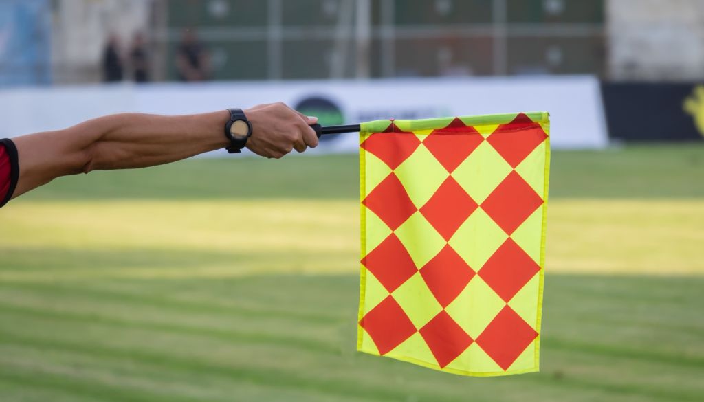 A person's hand holding a yellow and red checkered flag, used as a referee's signal in a sports event.