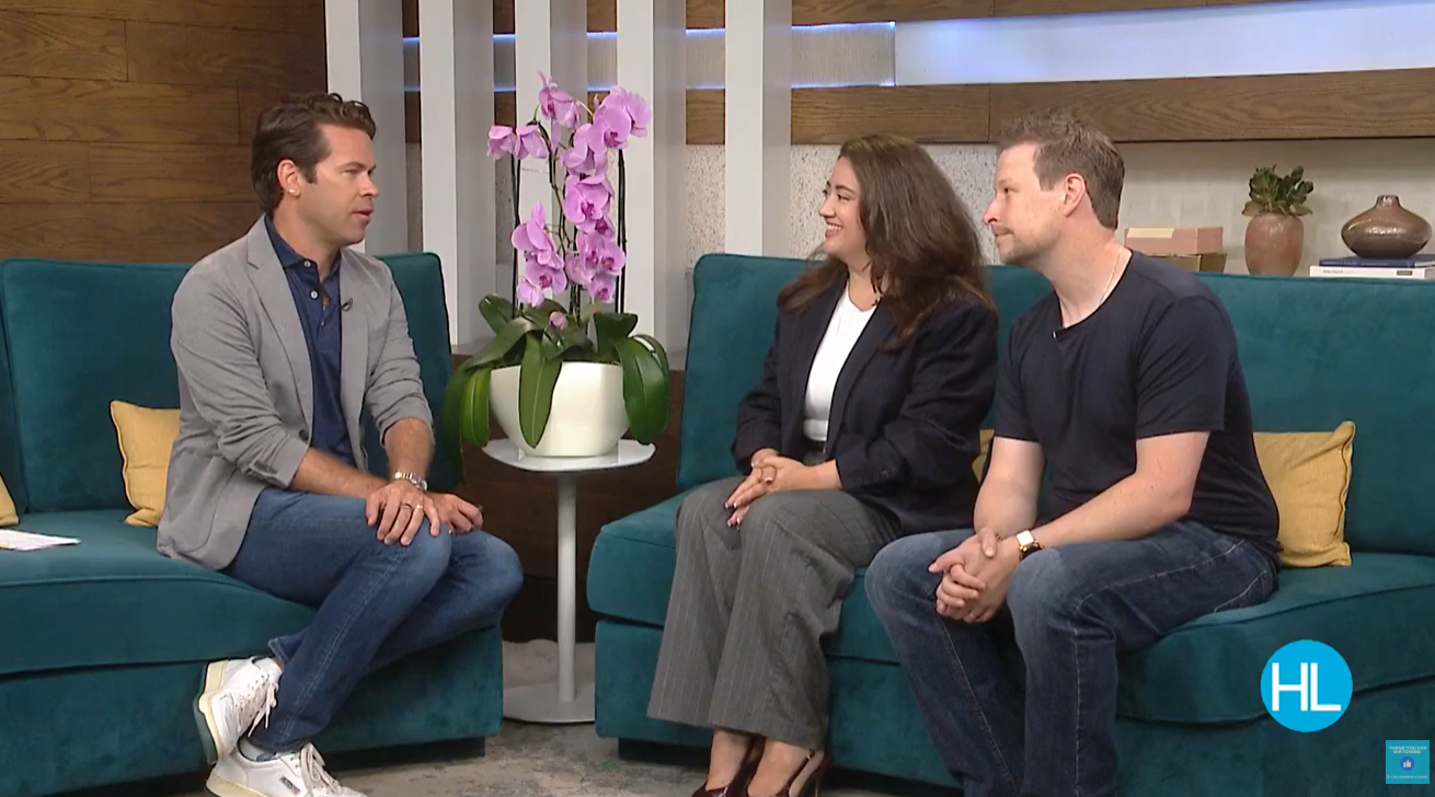 Three people sitting on a couch in a studio setting, with a potted orchid plant and other decor visible.